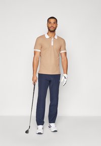 Caurspīdīgs polo krekls ar baltu apkakli un piedurknēm, apvienots ar tumši ziliem biksēm. Modelis tur golfa nūju un valkā baltas sporta kurpes. Gluda tekstūra.