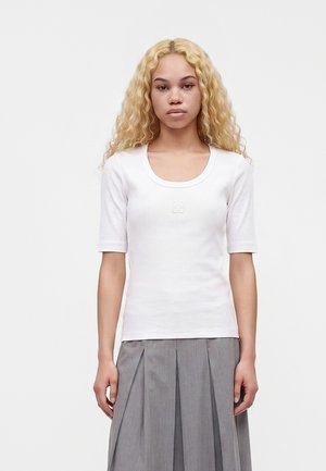 Giovane donna con capelli biondi ricci indossa una maglietta bianca a maniche corte e una gonna grigia plissettata, in piedi davanti a uno sfondo bianco uniforme.