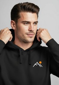 Schwarzer Hoodie mit einem Berggrafik und orangefarbener Sonne. Weicher Stoff, Kapuze mit Kordelzug, gerippte Bündchen und Saum für eine enganliegende Passform.