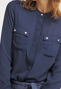 Camicia blu navy a maniche lunghe con botttoni, due tasche sul petto, dettagli dorati e design senza colletto. Tessuto liscio e vestibilità su misura.