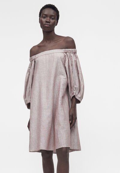 Γυναίκα που φοράει ένα φαρδύ ροζ φόρεμα έξω από τον ώμο με κάθετες λευκές ρίγες και μακριά φουσκωτά μανίκια σε απλό φόντο.