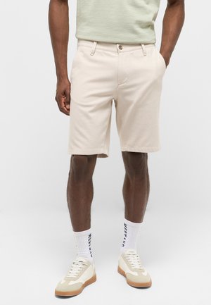 Mann trägt beige knielange Shorts, weiße Socken mit marineblauem Text, cremefarbene und weiße Sneakers, steht vor einem einfarbigen Hintergrund.