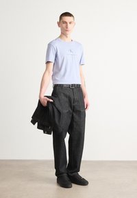 Camiseta de color púrpura claro con el logo "Calvin Klein Jeans", pantalones de mezclilla oscura, cinturón negro y zapatillas negras, sosteniendo una chaqueta de mezclilla negra.