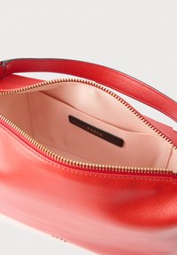 Punainen nahkalaukku, jossa on teksturoitu pinta, kullanvärinen vetoketjusuljenta ja sileä vaaleanpunainen sisävuori, jossa pieni tasku. Sisäpuolella näkyy brändi-läppä.
