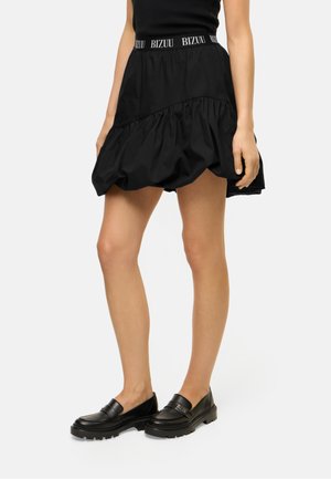 Vrouw met een zwarte gelaagde minirok met "BIZUU" op de tailleband en zwarte loafers, staat met haar armen ontspannen langs haar zij.