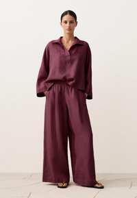 Nő áll, laza, hosszú ujjú szilvaszínű ingben és bő szárú nadrágban, fekete szandálokkal világos kövekből álló padlón, egyszínű háttér előtt.