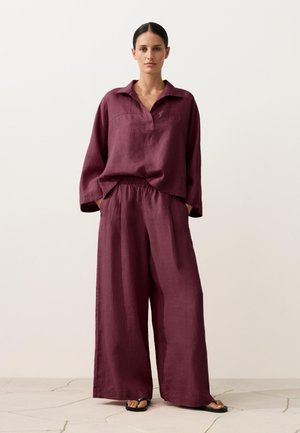 Vrouw staat in een losse, lange mouwen pruimenkleurige blouse en wijde broek met zwarte sandalen op een lichte stenen vloer en een effen achtergrond.