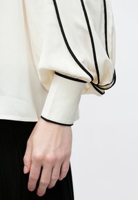 Hand and wrist in a long-sleeve white blouse with black trim on cuff and sleeve edges against a plain background.