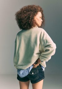 Lysegrå oversized fleece-sweatshirt lagdelt over en blåstribet skjorte, kombineret med mørke denimshorts. Krøllet hår synligt bagfra.