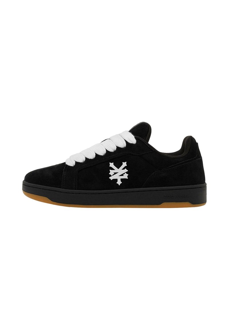 Svart semsket sneakers med hvite snører, med en hvit logo på siden, rund tå og gummisåle.