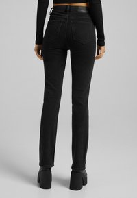 Jeans de mezclilla negra con un diseño ajustado, tiro medio, dos bolsillos traseros y detalles de costura. Combinados con botas de tacón grueso negras.