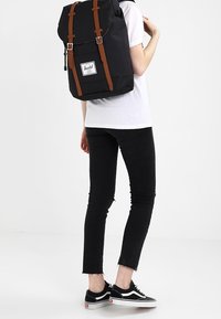 Sac à dos en tissu noir avec des sangles en cuir marron, fermeture roulée et patch de logo. Porté par une personne en chemise blanche et en jeans noirs.