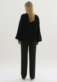 Svart överdimensionerad stickad tröja med en texturerad yta, vida ärmar och ribbat nederkant, i kombination med skräddarsydda svarta byxor. Neutral bakgrund.