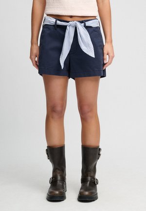 Vrouw in marineblauwe korte broek met een gestrikte gestreepte riem en donkerbruine leren laarzen tot halverwege de kuit, staand tegen een effen achtergrond.