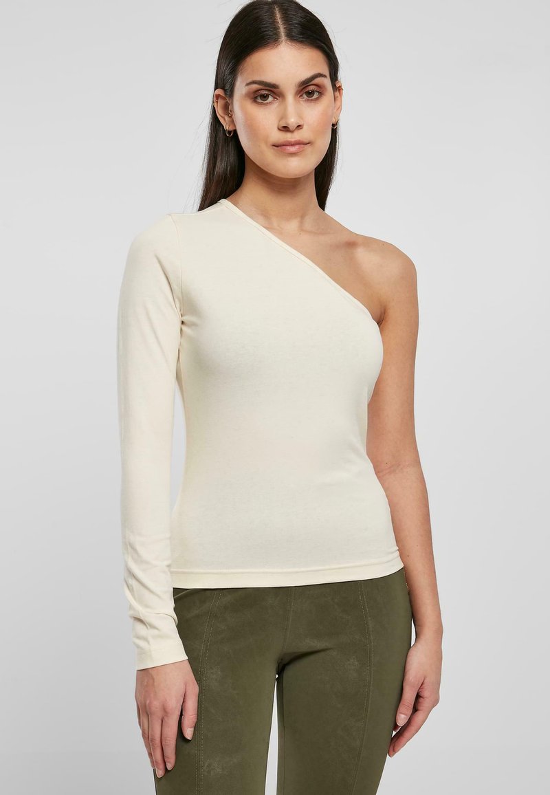 Asymmetriska beige långärmade topp med en axel bar, figursydd design, lättviktsmaterial och mjuk textur, i kombination med olivgröna byxor.