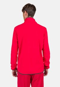 Red fleece jacket with a high collar, long sleeves, and a curved hem. Material appears soft; features minimal stitching and no visible logos.