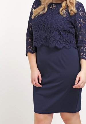 Dame draagt een marineblauw kanten top met driekwart mouwen en een bijpassende knie-lange rok, met een broche op de borst.