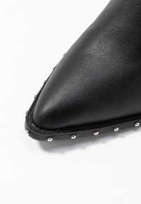 Scarpa di pelle nera con punta appuntita, caratterizzata da dettagli in metallo argentato lungo la suola e una finitura texturizzata.