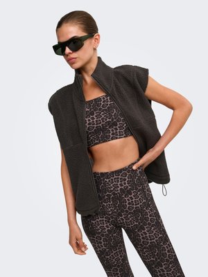 Femme portant un gilet polaire sans manches noir par-dessus un soutien-gorge de sport et des leggings à imprimé léopard, avec des lunettes de soleil, sur fond uni.