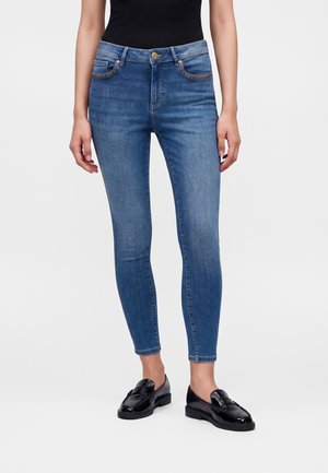 Kvinde iført blå skinny jeans og sorte loafers, stående med hænderne afslappet ned langs siderne mod en hvid baggrund.
