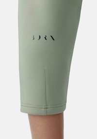 Pantaloni sportivi verdi realizzati in tessuto liscio e elastico. Presentano un logo nero sul lato sinistro e un orlo rifinito con una cucitura pulita.