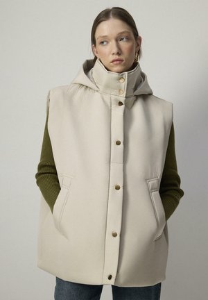 Gilet rembourré beige avec capuche, boutons-pression dorés, poches latérales et manches longues vertes en dessous. Texture lisse, coupe oversize.