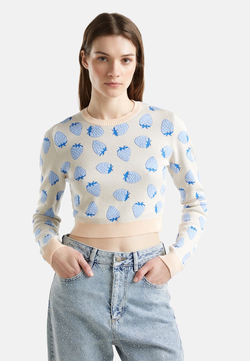 Sweter na szydełku w kolorze kremowym z niebieskimi wzorami truskawek, prążkowany dekolt i mankiety, krótki fason, połączony z jasnymi dżinsami.