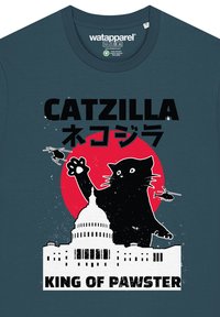 T-shirt bleu foncé avec un graphique représentant un grand chat noir devant un cercle rouge, un bâtiment blanc et des hélicoptères. Le texte dit "CATZILLA ROI DE PAWSTER."