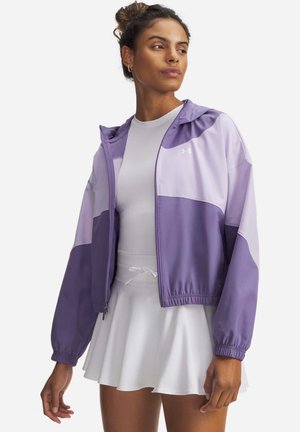 Donna che indossa una giacca viola a blocchi con cerniera sopra un vestito sportivo bianco con cintura annodata, guardando di lato.