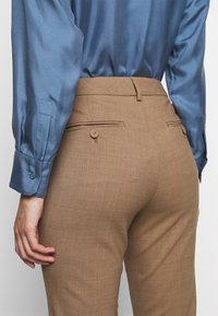 Personne portant un pantalon marron taille haute avec des poches arrière boutonnées et une chemise en satin bleu à manches longues rentrée, main détendue sur le côté.