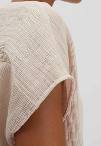 Ljust rosa strukturerad tyg med vertikala ränder, med en lös, draperad ärmdesign. Detalj av tygets vågiga yta är synlig.