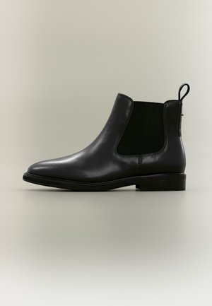 Bottine Chelsea noire en cuir avec panneau latéral élastique et languette sur fond neutre.