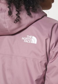 Ljust rosa vattentät jacka med huva, med en vit logotyp av "The North Face" på den övre delen av ryggen, slät textur och minimal sömnad.