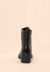 Bota de cuero negra hasta el tobillo con tacón cuadrado, cremallera lateral y tirador, mostrada desde la parte trasera sobre un fondo beige.