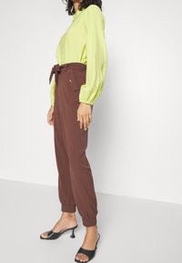 Kvinna iklädd en ljusgrön långärmad blus, högmidjade bruna papperspåsebyxor med knytbälte och svarta sandaler med öppen tå och kitten heels.