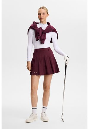 Vrouw draagt een witte top met lange mouwen, een bordeaux geplooide rok, een bordeaux trui over de schouders, witte handschoen, en houdt een golfclub vast.
