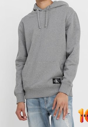 Osoba ubrana w szarą bluzę z kapturem i kieszenią na przodzie oraz z naszywką Calvin Klein na dole, w połączeniu z spranymi jasnoniebieskimi dżinsami.