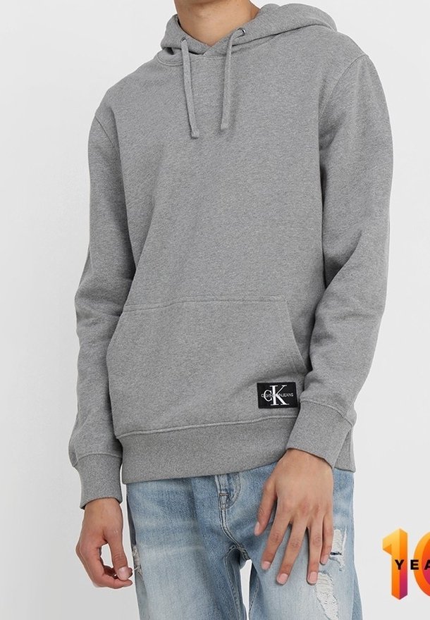 Personne portant un sweat à capuche gris avec poche kangourou à l'avant et un patch Calvin Klein sur le bas, associé à un jean bleu clair délavé.