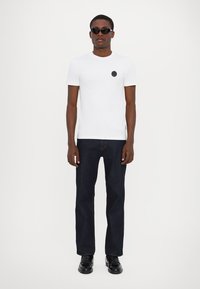 Hvid t-shirt med korte ærmer og en sort cirkulær lap, parret med mørkeblå lige bukser og sorte blanke sko, accessoriseret med solbriller.