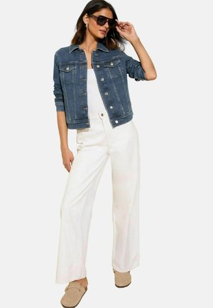 Giacca di denim blu scuro, chiusura a bottoni, due tasche sul petto. Pantaloni bianchi a gamba larga, scarpe beige senza stringhe e occhiali da sole neri oversize.