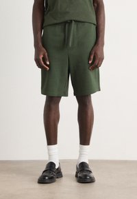 Grønne bomuldsblandingsshorts med snoretræk i taljen, sideslids ved kanten og afslappet pasform, parret med sorte loafers og hvide sokker.
