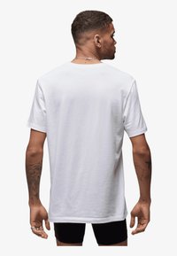 Weißes Baumwoll-T-Shirt mit kurzen Ärmeln, lockerer Passform. Hat ein schlichtes Rückendesign und eine glatte Textur, getragen mit schwarzen Shorts.