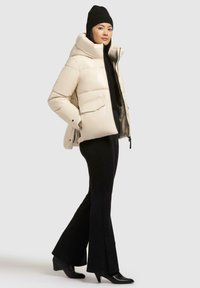 Giacca a vento color crema con colletto alto, caratterizzata da ampie tasche frontali e chiusura con zip. Abbinata a pantaloni neri a campana e stivali con tacco.