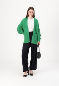 Grønn, fluffy cardigan med åpen foran, iført over en hvit topp og svarte stripete vide bukser. Svarte spissede hæler og veske.