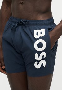 Man met marineblauwe zwembroek met witte "BOSS" tekst, elastische tailleband en trekkoord, hand in rechterzak, tegen een effen achtergrond.