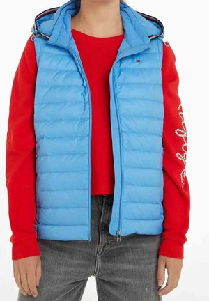 Henkilö, joka käyttää vaaleansinistä toppaliivihattua punaisen pitkähihaisen paidan päällä, jossa on valkoista tekstiä vasemmassa hihassa, ja tummanharmaat farkut.