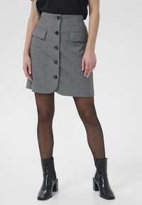 Jupe grise rayée avec une taille ajustée, comportant quatre boutons noirs, deux poches avant et un ourlet légèrement évasé. Portée avec des bottes à talons noires.