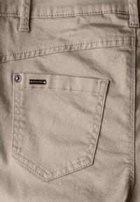 Pantalons beiges avec une finition texturée, dotés d'une poche arrière avec un bouton-pression en métal argenté et d'une petite étiquette en métal de marque. Les détails de couture ajoutent de l'intérêt.