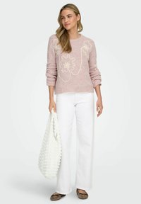 Lys rosa strikket genser med hvit blomsterbroderi, kombinert med hvite vidde bukser og en strukturert hvit veske. Brune leppa fullfører antrekket.