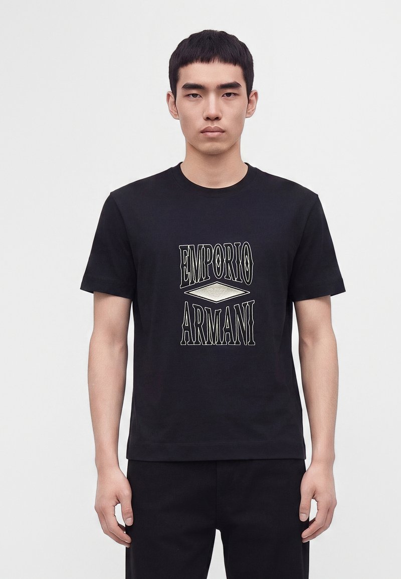 Νεαρός άνδρας που φοράει μαύρο T-shirt με κείμενο και λογότυπο "EMPORIO ARMANI" στο μπροστινό μέρος, στέκεται μπροστά σε λευκό φόντο.